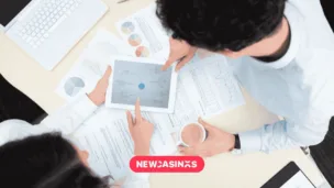 UKGC Deepens Consumer Engagement with New Research Partnerships An overhead shot of two people conferring over a tablet screen showing a plan for business growth alongside papers, alongside a 'NEWCASINOS' logo in pink.