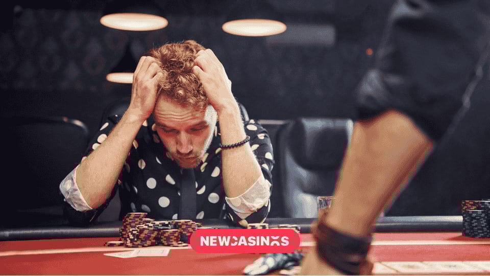A distressed man sits at a casino table with his hands on his head, surrounded by poker chips, conveying a sense of gambling loss and emotional struggle. The logo "NewCasinos" is visible in the foreground.