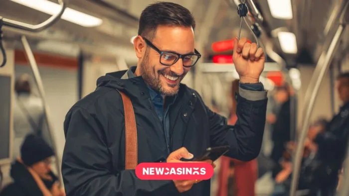 A man with glasses and backpack standing on a train or underground. He is holding a phone and looking at it whilst smiling, the New Casinos logo in the foreground.