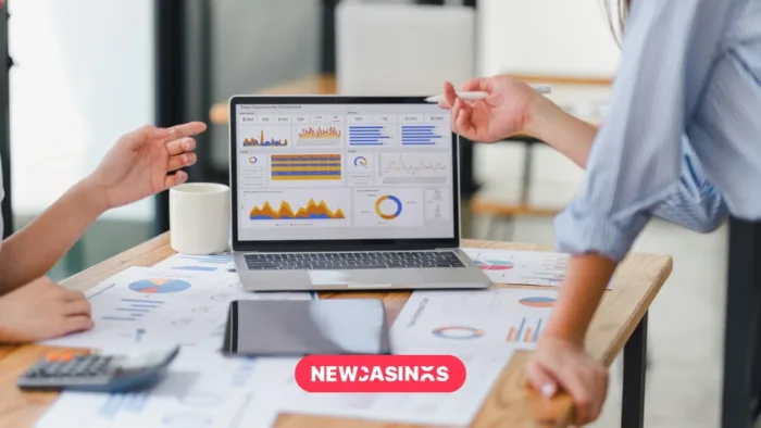 Screen showing graphs and other graphics with papers across the table and two business people talking.